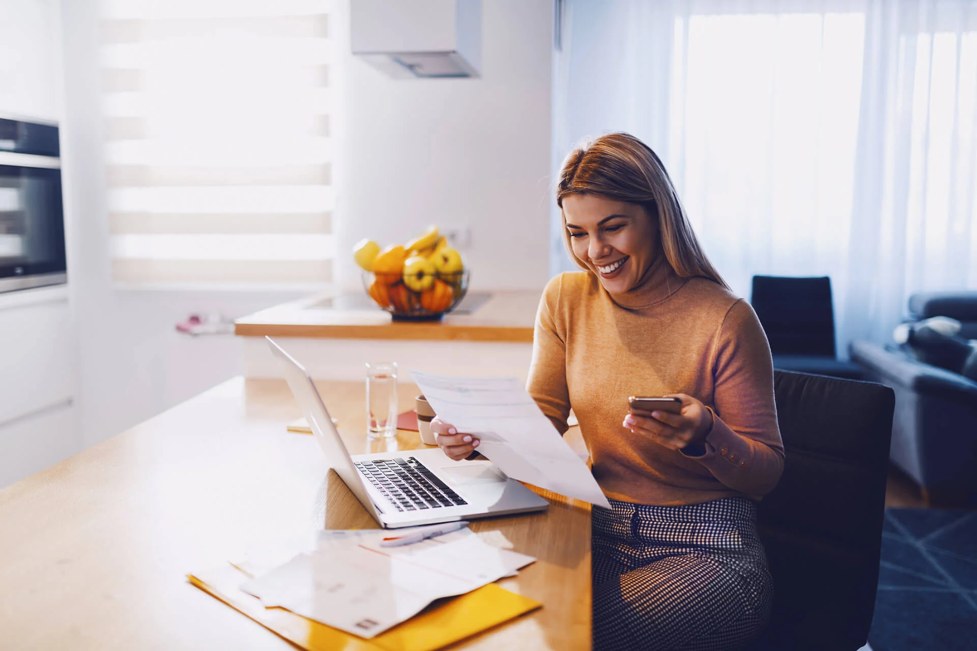 A woman smiling at her kitchen table while reviewing her paperwork with a laptop and phone